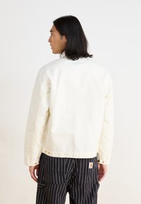 Witte katoenen jas met een relaxte pasvorm, voorzien van een knoopsluiting en minimale details, gedragen over een donkere gestreepte broek. Achteraanzicht.
