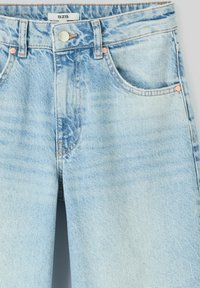 Jean en denim bleu clair avec poches avant, rivets en cuivre, passants de ceinture et fermeture à bouton argenté, affichant une étiquette de marque BZB.