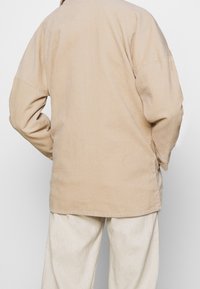 Beige kordjacka med lös passform, med vida ärmar och en texturerad yta. Baksidan har en enkel design utan utsmyckningar.