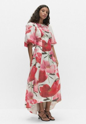 Vrouw met lang krullend haar, gekleed in een witte high-low jurk met grote rode en roze bloemenprints en zwarte sandalen met bandjes.