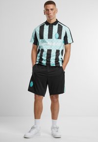 Polo shirt met lichtblauwe en zwarte strepen, met een geometrisch patroon en logo; gecombineerd met zwarte shorts en witte sneakers.