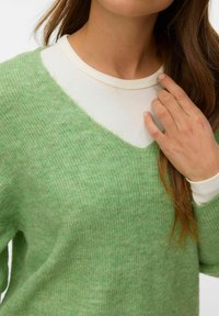 Femme portant un t-shirt blanc côtelé sous un pull en maille vert, touchant l'encolure avec sa main droite.