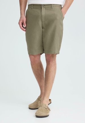 Mand iført olivengrønne shorts, en hvid skjorte og tanfarvede slip-on ruskindssko, stående med hænderne i lommerne.