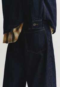 Jeans jakna in hlače v temno modri barvi z rumenim šivanjem, gumbnim detajlom in pod njo videno karo srajco.