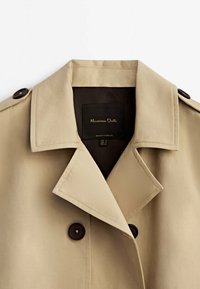 Beżowy płaszcz trench z szerokim kołnierzem, z ciemnymi guzikami i gładką teksturą. Metka wewnątrz mówi "Massimo Dutti."