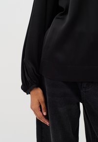 Blusa nera a maniche lunghe con una texture liscia e polsini arricciati, abbinata a jeans scuri. La blusa ha una vestibilità comoda e una leggera lucentezza.