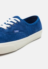 Baskets Vans en daim bleu avec lacets blancs et semelle en caoutchouc écrue, présentées en angle pour montrer les détails latéraux et avant sur un fond uni.