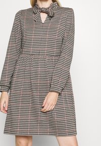 Robe à motifs noir et blanc avec un col en nœud papillon, des manches longues et une taille élastique. Présente une découpe en trou de clé et une jupe évasée.