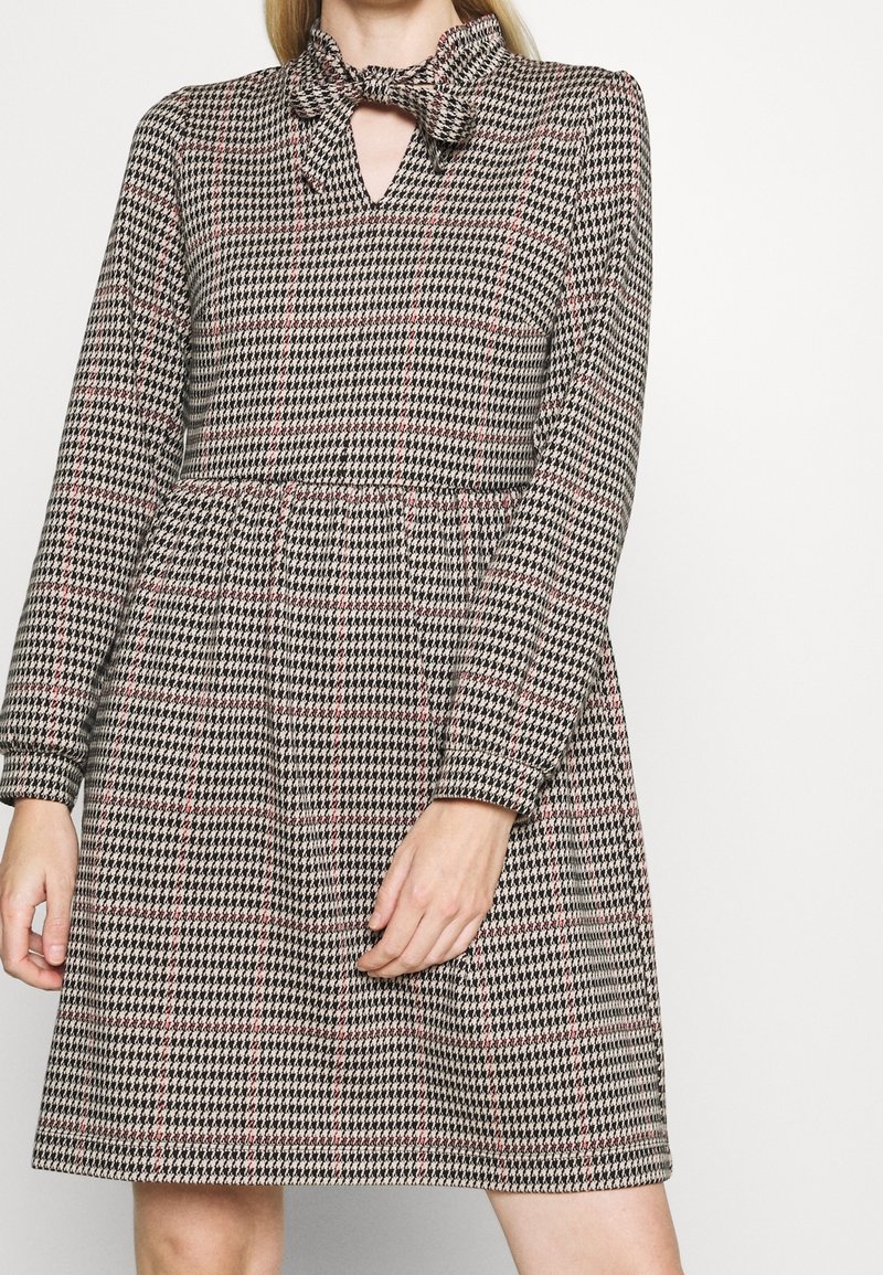 Robe à motifs noir et blanc avec un col en nœud papillon, des manches longues et une taille élastique. Présente une découpe en trou de clé et une jupe évasée.