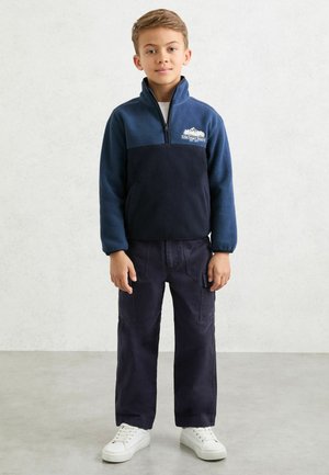 Tumesinise tõmblukuga fliisjakk kontrastse tumeda sinise paneeliga, millel on mäelogo. Kantud tumedate cargo-pükste ja valgete tossudega.