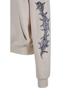 Beige hættetrøje med et sort torn og rose broderet design langs venstre ærme. Indeholder en frontlomme og ribbede manchetter.