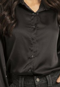 Chemise en satin noir à boutonner avec col, dotée de six boutons et d'une texture lisse, associée à un pantalon en denim noir.