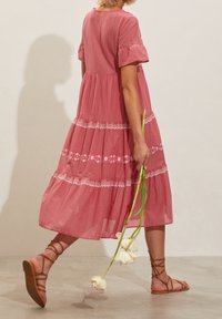 Vaaleanpunainen, kerroksellinen mekko lyhyillä hihoilla ja valkoisilla kukka-ompeluilla. Yhdistetty ruskeiden remmi-sandaalien kanssa, pidellen valkoisia tulppaaneja.