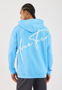 Uomo con capelli intrecciati che indossa un hoodie azzurro chiaro con la scritta in corsivo bianco "Your Turns" sulla schiena, in piedi contro uno sfondo semplice.