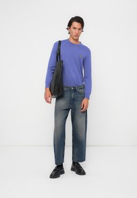 Lila Langarmpullover mit kleinem gelben Logo, kombiniert mit hellblauen Weitbeinhosen und schwarzen Schuhen. Hält eine schwarze Tragetasche.