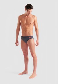 Bañador tipo slip de color gris oscuro con un logo naranja de "arena" en la parte frontal, con un corte bajo y textura suave. El modelo está descalzo.