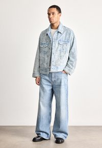 Man wearing light blue textured jacket, white shirt, wide-leg faded jeans, and black shoes standing against plain background.