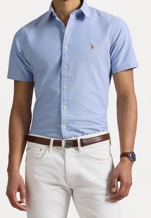 Man wearing a light blue short-sleeve button-up shirt with a small logo, white pants, brown leather belt, and wristwatch with a brown strap.