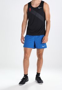Camiseta sin mangas negra de Nike con logo rojo y franjas diagonales; pantalones cortos azules; zapatillas de correr negras con acentos blancos; calcetines negros lisos.