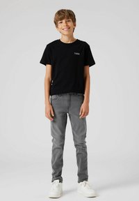 Maglietta nera in cotone con logo "TWSN", abbinata a jeans grigi a vita bassa e sneakers bianche, indossata da un bambino in piedi davanti a uno sfondo neutro.