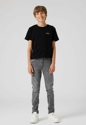 Mustast puuvillast T-särk "TWSN" logoga, millele on lisatud hallid kitsad teksapüksid ja valged tossud, mida kannab laps, kes seisab neutraalse tausta ees.
