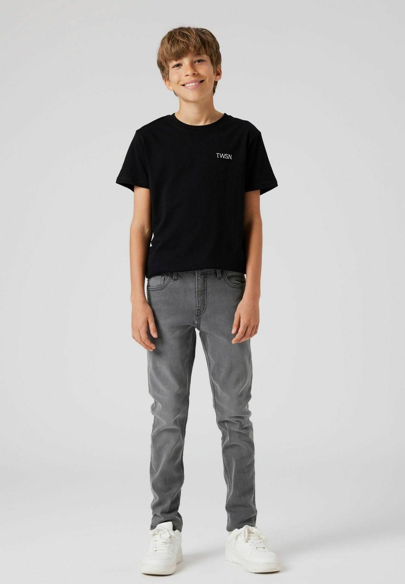 Maglietta nera in cotone con logo "TWSN", abbinata a jeans grigi a vita bassa e sneakers bianche, indossata da un bambino in piedi davanti a uno sfondo neutro.
