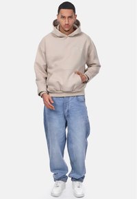 Beige hooded sweatshirt with a kangaroo pocket, paired with light blue baggy jeans and white sneakers. Smooth fabric texture.