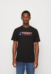Černé bavlněné tričko s výrazným červeným a bílým logem "TOMMY JEANS", s grafikou americké vlajky, krátkými rukávy a kulatým výstřihem.