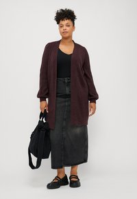 Cardigan in magenta con apertura frontale, maniche lunghe e voluminose, e finitura testurizzata, indossato sopra un top nero e abbinato a una borsa nera.