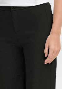 Black trousers made of a textured fabric, with a slim fit, zip and button closure, and side pockets. Hand resting on the fabric for scale.