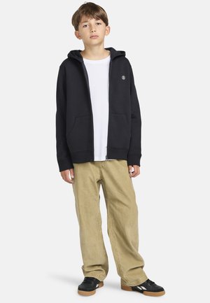 Svart zip-up hoodie med en framficka, bärs över en vit T-shirt, ihop med beige vida byxor och svarta sneakers.