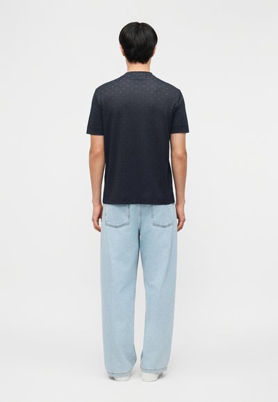 Άνδρας που φοράει μαύρο κοντομάνικο πουκάμισο με μικρά σχέδια και ανοιχτό μπλε παντελόνι με φαρδιά μπατζάκια, στέκεται με την πλάτη γυρισμένη σε λευκό φόντο.