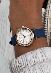 Ronde horloge met een roségouden lunette en een wit wijzerplaat. Blauwe siliconen band. Voorzien van kristallen accenten bij de uurmarkeringen en merkje.