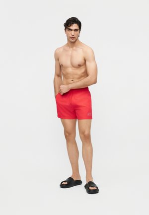 Hombre sin camiseta, con pantalones cortos de baño rojos brillantes y sandalias negras tipo chancla, de pie con una mano en el bolsillo contra un fondo blanco.