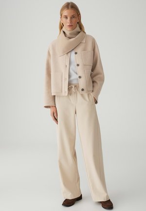 Veste beige en mélange de laine avec grandes poches, fermeture à boutons et large col, associée à un pantalon à jambes larges crème et des chaussures marron.