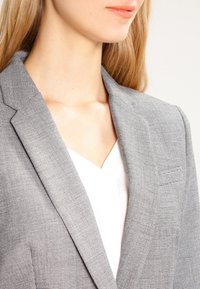 Gray patterned blazer with a notched collar, structured shoulders, and subtle texture, paired with a white top beneath.