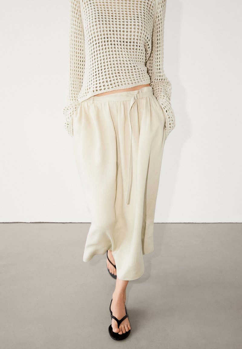 Personne portant un pantalon large crème avec une ceinture à nouer, un pull en maille ample et des sandales noires à bout ouvert marchant à l’intérieur.