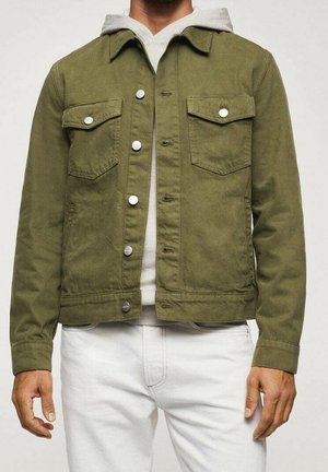 Mężczyzna w oliwkowozielonej kurtce zapinanej na guziki, na jasnoszarej bluzie z kapturem i białych spodniach, stojący na tle jednolitego jasnego tła.