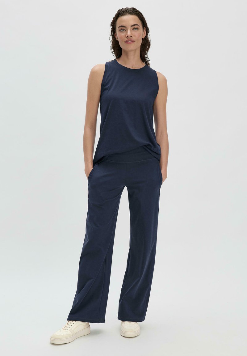 Marineblaues ärmelloses Oberteil kombiniert mit locker sitzenden marineblauen Hosen, beide aus einem weichen Stoff. Das Model trägt cremefarbene Turnschuhe.