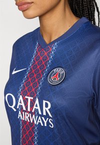 Blå Nike-tröja med ett röd, vit och blått geometriskt mönster; inkluderar texten "QATAR AIRWAYS" och en cirkulär Paris Saint-Germain-logotyp.
