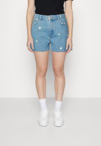 Jeansshorts in Hellblau, mit bestickten weißen Gänseblümchen verziert. Ausgestattet mit einem umgeschlagenen Saum und vorderer Knopfleiste. Kombiniert mit weißen Turnschuhen.