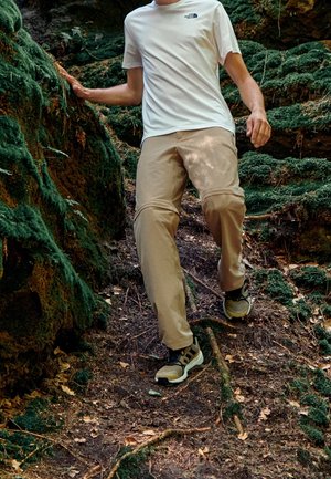 Person trägt beige Wanderhose und braune Schuhe, geht einen moosbedeckten, unebenen Waldweg hinunter und stützt sich mit der Hand auf einen Felsen ab.