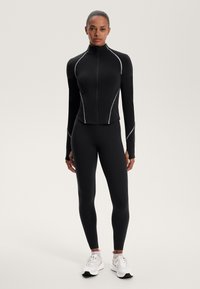 Fekete sportdzseki cipzárral és hosszú ujjakkal, fekete leggingsszel és fehér sportcipővel párosítva. Kontrasztként fehér varrással rendelkezik.