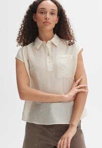 Helle beige Kurzarmbluse aus strukturiertem Stoff mit einem Kragen, durchgehender Knopfleiste und einer einzelnen Brusttasche.