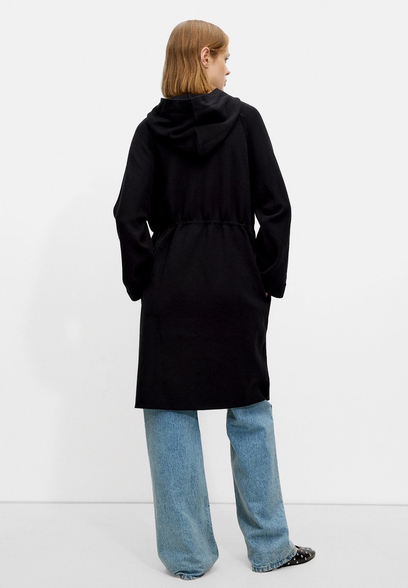 Manteau long noir à capuche avec une taille marquée, présentant de longues manches et une texture lisse, associé à un jean à jambes larges bleu clair.