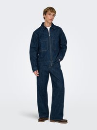 Mørkeblå denimjakke med to brystlommer, kombinert med matchende bukser med løs passform. Antrekket har glidelåslukking og vide ben.