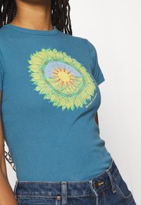 Blue cotton t-shirt featuring a vibrant sunflower and sun graphic, with green accents and text "Mother Earth" near the hem.