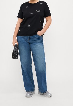 Person wearing black T-shirt with small handbag prints and text, blue jeans, silver sneakers, holding black handbag with metal eyelets.