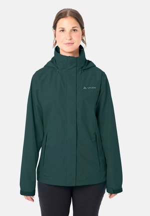 Frau trägt dunkelgrüne wasserfeste Jacke mit hohem Kragen und Kapuze, steht vor einem schlichten hellen Hintergrund.