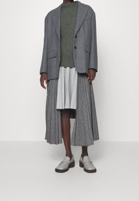 Osoba w zielonym swetrze z dzianiny, szarym oversize'owym żakiecie, plisowanej dwutonowej szarej spódnicy midi oraz szarych platformowych mokasynach.